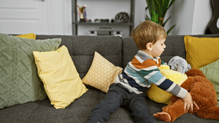 Blond toddler boy relaxing on a sofa with colorful pillows and plush toys in a cozy living room.
