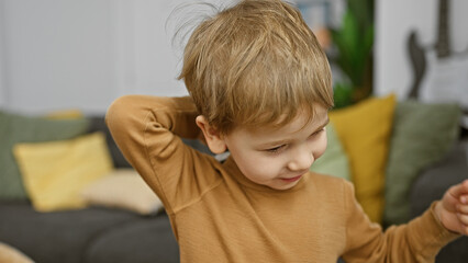 A blonde toddler boy in a living room, wearing a brown shirt with a cozy couch and pillows in the background.