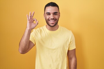 Young hispanic man standing over yellow background smiling positive doing ok sign with hand and fingers. successful expression.