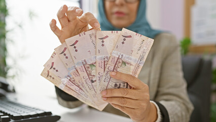 A woman in a hijab counts saudi riyal bills at her office, suggesting a business transaction or...