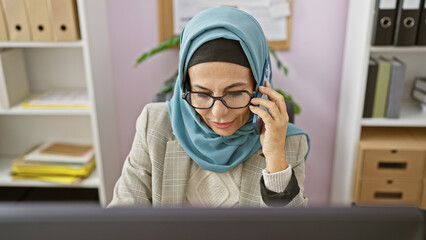 A professional woman wearing hijab and glasses speaking on the phone in a modern office setting.