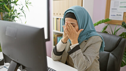 A stressed middle-aged woman in hijab feeling overwhelmed at her office workstation, eyes covered,...