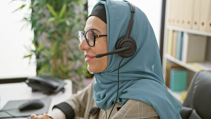 A professional woman wearing a hijab and headset working in a modern office environment. - obrazy, fototapety, plakaty