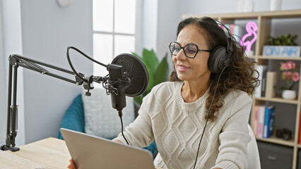 A smiling mature hispanic woman wearing headphones speaks into a studio microphone while reading...