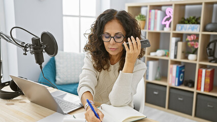 Hispanic woman listening voice message indoors at podcast studio with laptop and microphone