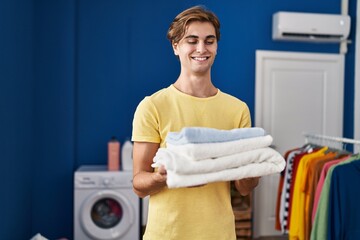 Young caucasian man smiling confident holding folded towels at laundry room