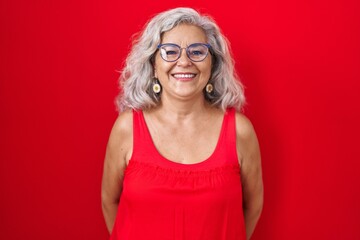 Middle age woman with grey hair standing over red background with a happy and cool smile on face....