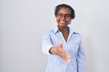 African woman with dreadlocks standing over white background wearing glasses smiling friendly offering handshake as greeting and welcoming. successful business. - obrazy, fototapety, plakaty
