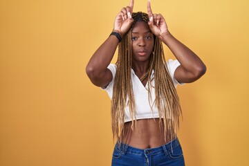 African american woman with braided hair standing over yellow background doing funny gesture with...