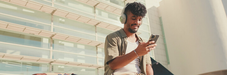 Young male student dressed in an olive color shirt sits outside next to the university, listens to music on headphones, selects tracks on his phone, Panorama