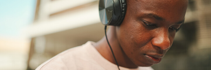 Close-up portrait of young african student sitting outside of university wearing headphones,...