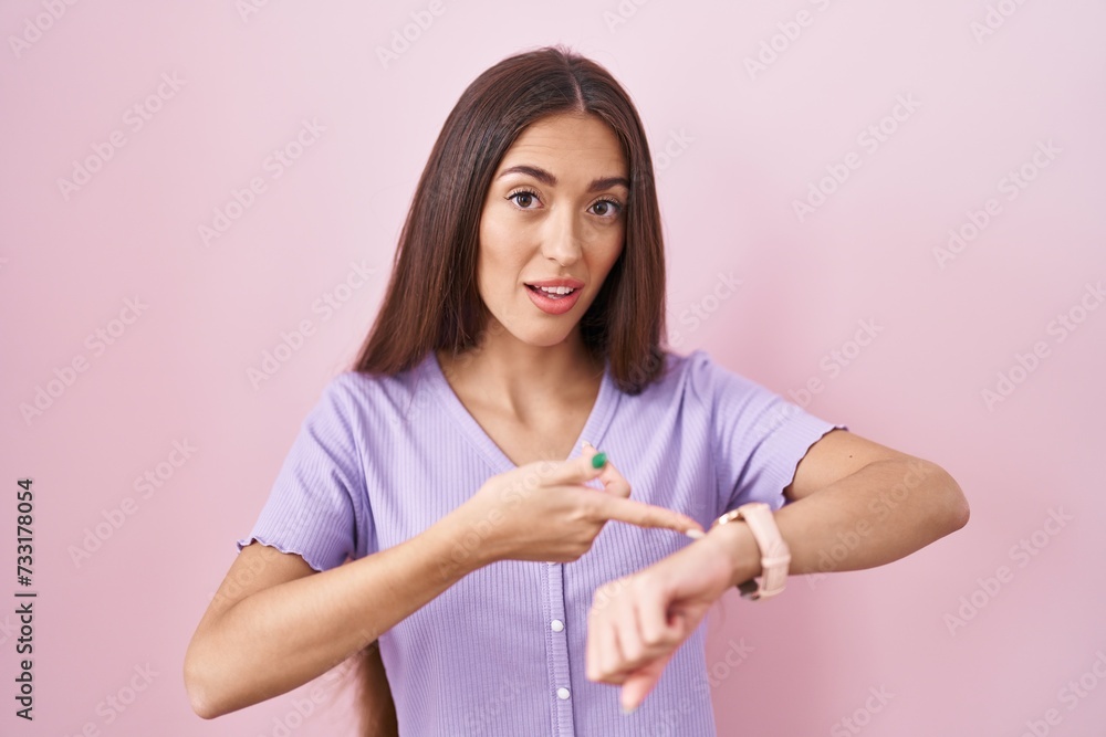 Poster young hispanic woman with long hair standing over pink background in hurry pointing to watch time, i