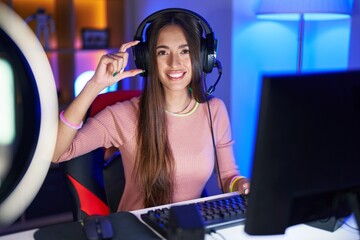 Young hispanic woman playing video games smiling and confident gesturing with hand doing small size sign with fingers looking and the camera. measure concept.