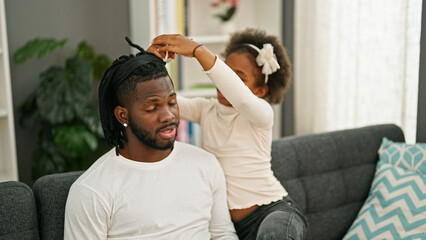 African american father and daughter sitting on sofa playing with hair doing pigtail at home