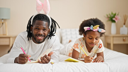 African american father and daughter wearing funny diadem drawing on notebook lying on bed at bedroom - obrazy, fototapety, plakaty