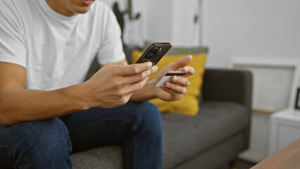 Young hispanic man using a credit card and smartphone for online shopping in a cozy home interior.