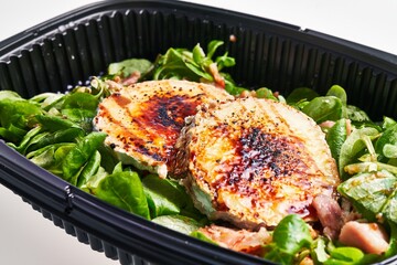 Delicious grilled chicken breast atop a fresh spinach salad, served in a black takeout container, depicts a healthy meal.