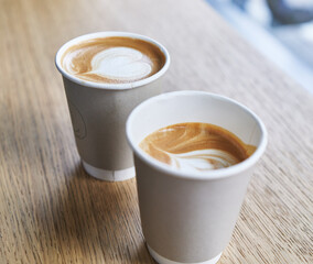 Two takeaway paper cups of cappuccino with heart-shaped latte art sitting on a wooden table.