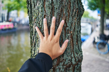 A person touches a textured tree trunk by a canal, suggesting connection to nature in an urban...