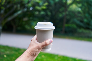 Man's hand presenting takeaway coffee in a park setting, depicting a quick caffeine fix in a...