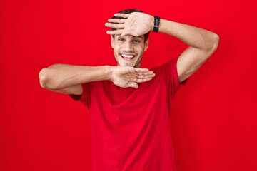 Young hispanic man standing over red background smiling cheerful playing peek a boo with hands showing face. surprised and exited