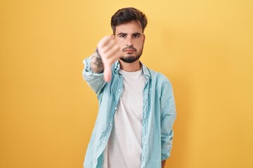 Young hispanic man with tattoos standing over yellow background looking unhappy and angry showing...
