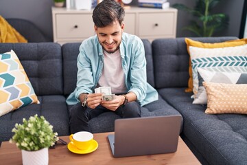 Young hispanic man using laptop counting dollars at home
