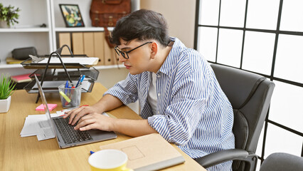 Professional young man typing on laptop at his organized office desk with window backdrop