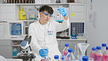 A young man inspects a flask in a laboratory full of equipment like beakers and a microscope.