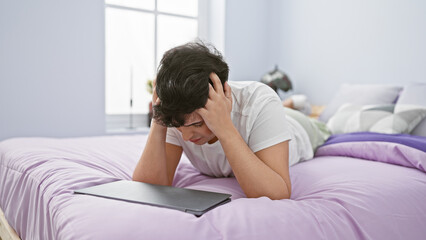 Distressed young man with laptop in a modern bedroom conveying frustration or headache.
