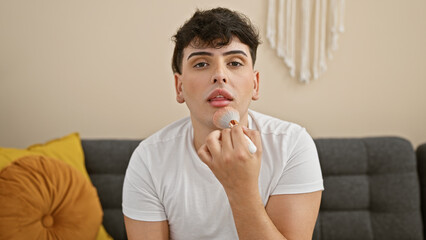 A handsome young man applies makeup with a brush in a modern living room, showcasing gender fluidity and self-care at home.