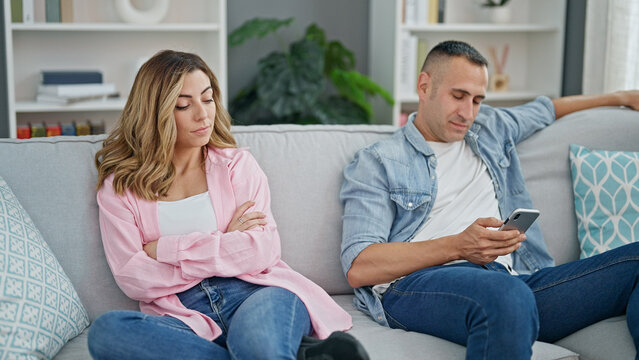 Man and woman couple using smartphone sitting on sofa looking upset at home