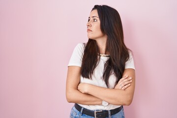 Young brunette woman standing over pink background looking to the side with arms crossed convinced...