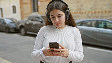 A young hispanic woman wearing headphones enjoys music on her smartphone in a bustling city street.