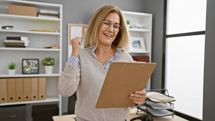 A joyful mature woman celebrates success with a clipboard in a modern office setting, indicating...