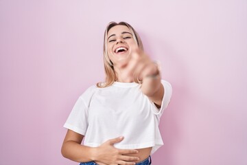 Young blonde woman standing over pink background laughing at you, pointing finger to the camera...