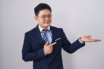 Young asian man wearing business suit and tie amazed and smiling to the camera while presenting with hand and pointing with finger.