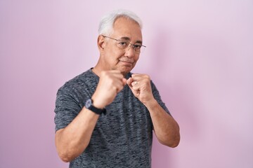 Middle age man with grey hair standing over pink background ready to fight with fist defense gesture, angry and upset face, afraid of problem