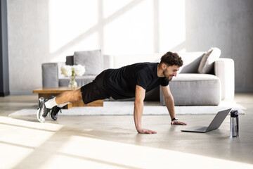 Happy man in sportswear doing push-ups, using laptop, having online fitness class from home