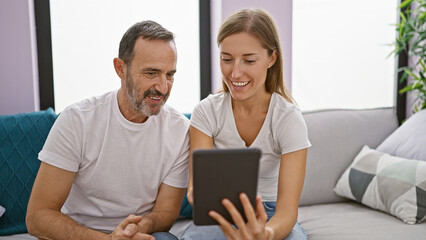Fototapeta premium Father and daughter share a jovial moment, confidently smiling while sitting together on the homely sofa, fully engrossed in their touchpad, enjoying their relaxed online lifestyle.