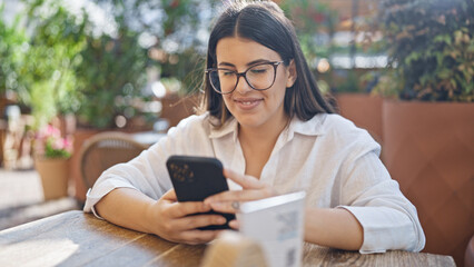 Fototapeta premium Young beautiful hispanic woman smiling happy using smartphone sitting on the table at bar terrace