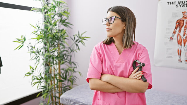 Confident hispanic woman in pink scrubs standing with arms crossed in a clinic interior beside a reflex hammer and anatomical chart.