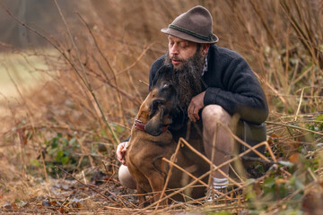 A bavarian man wearing a traditional folk costume interacting with his bavarian mountain dog nearby