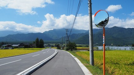 Road in the middle of grass fields, with a road safety convex mirror, with Japanese Alps mountains, blue sky and white clouds, in Azumino, Nagano Prefecture, Japan (Japanese words being driving alerts