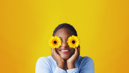  Playful brunette african woman covering her eyes with colorful flowers.