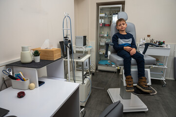 Fototapeta premium little emotional boy, fair-haired teenager, sitting in the otolaryngologist's office, waiting for the doctor for an examination, but smiling. Physical examination