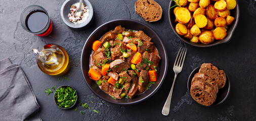 Beef meat and vegetables stew in black bowl with roasted baby potatoes. Dark background. Top view. - obrazy, fototapety, plakaty