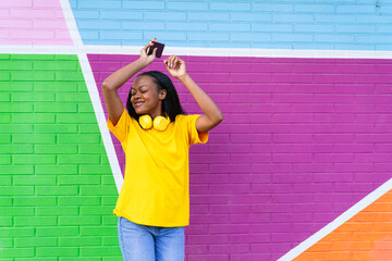 African woman in yellow feels the rhythm, dancing with phone in hand against a diagonally striped colorful wall.