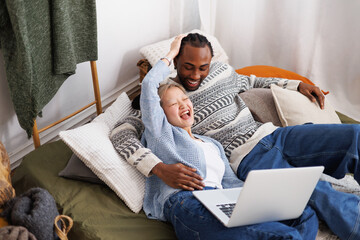 Cheerful young multiethnic couple relaxing near laptop on bed at home