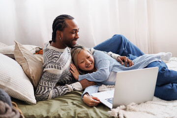 Positive young diverse couple looking at laptop while relaxing on bed - Powered by Adobe